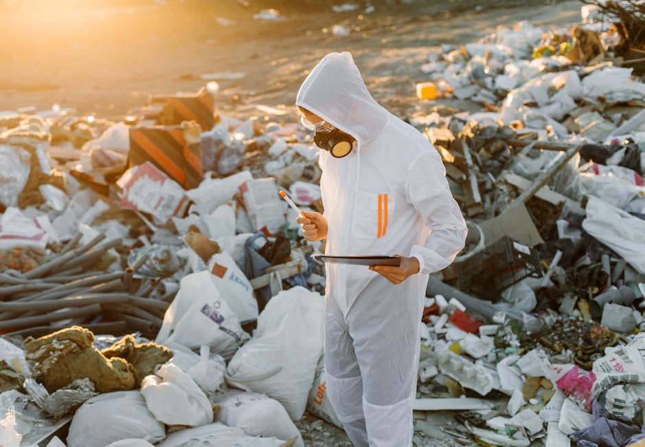 A person dressed in white protective coveralls wearing goggles and a respirator mask, holding a bright yellow container, standing outdoors in a landfill site filled with piles of mixed waste and debri