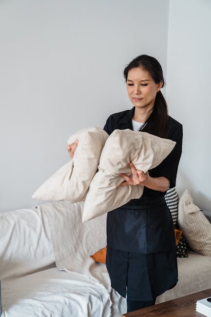 A professional cleaner, standing in a well-lit living room with white walls, holds two beige cushions in her hands, demonstrating domestic upholstery cleaning. The sofa, covered with a white slipcover, is situated against the wall and appears clean and tidy. In the background, decorative pillows, including a black and white patterned cushion and an orange cushion, are arranged on the sofa. The room features a wooden table visible in the foreground, and natural light fills the space, emphasizing the cleanliness and maintenance of the upholstery. The image reflects thorough surface cleaning and hygiene practices undertaken by Carpet Cleaning W10 in a residential setting, aligning with their specialist upholstery cleaning service in Notting Hill, W10.