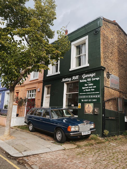 Exterior view of the Nottingham Hill Garage, a two-story building with a dark green facade and a large window on the ground floor, located in a residential area of Notting Hill. A grey vintage car is 